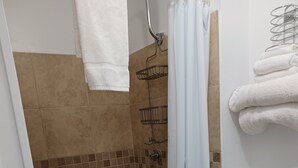 Un área de ducha con cortina blanca, una toalla blanca colgada en la pared y un estante con artículos de aseo.