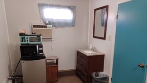 Una kitchenette compacta con microondas, tostadora y refrigerador. Hay un pequeño armario de madera con un lavamanos y un espejo encima. Una ventana con cortinas traslúcidas proporciona luz natural, y se ve una puerta azul.