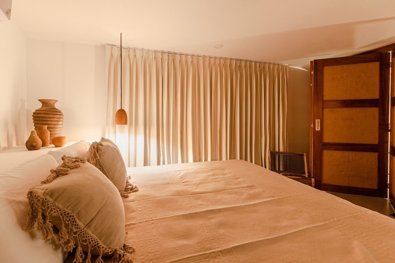 A bedroom with a large bed, beige curtains, a wooden door, and a vase on the nightstand.