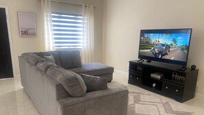 Un salon avec un canapé d’angle gris, un téléviseur à écran plat posé sur un meuble noir et un tapis à motifs.