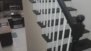 Un escalier avec des balustrades noires et blanches, menant à un espace cuisine doté de meubles sombres et d’appareils électroménagers en acier inoxydable.