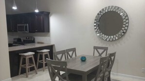 Un coin repas avec une table et des chaises en bois, un miroir rond accroché au mur, et une cuisine en arrière-plan.