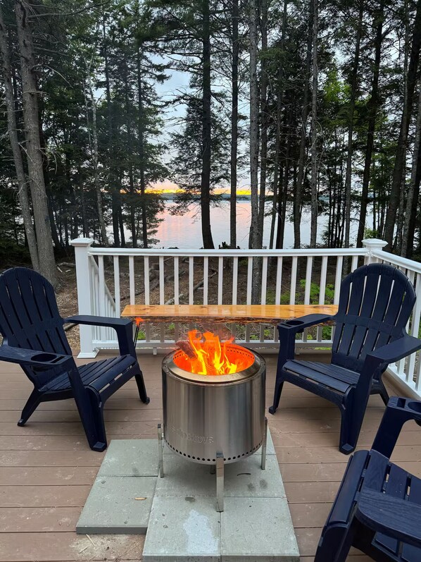A deck with a fire pit, two Adirondack chairs, and a bench overlooking a lake with trees.