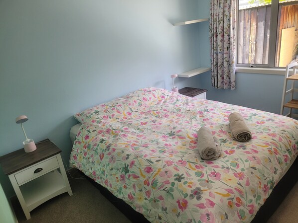 A bedroom with a floral bedspread, a bedside lamp, a small nightstand, and a window with curtains.