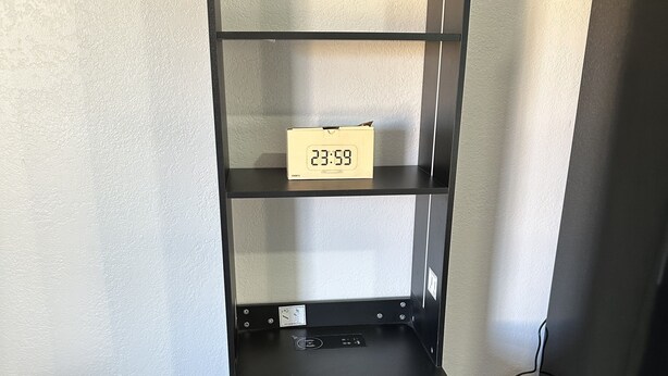 Una estantería metálica negra con un reloj digital que marca las 23:59, un cajón pequeño con tirador de latón y un suelo de madera.