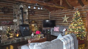 L’intérieur chaleureux d’un chalet en bois, avec une cheminée en pierre, un téléviseur et deux sapins de Noël décorés.