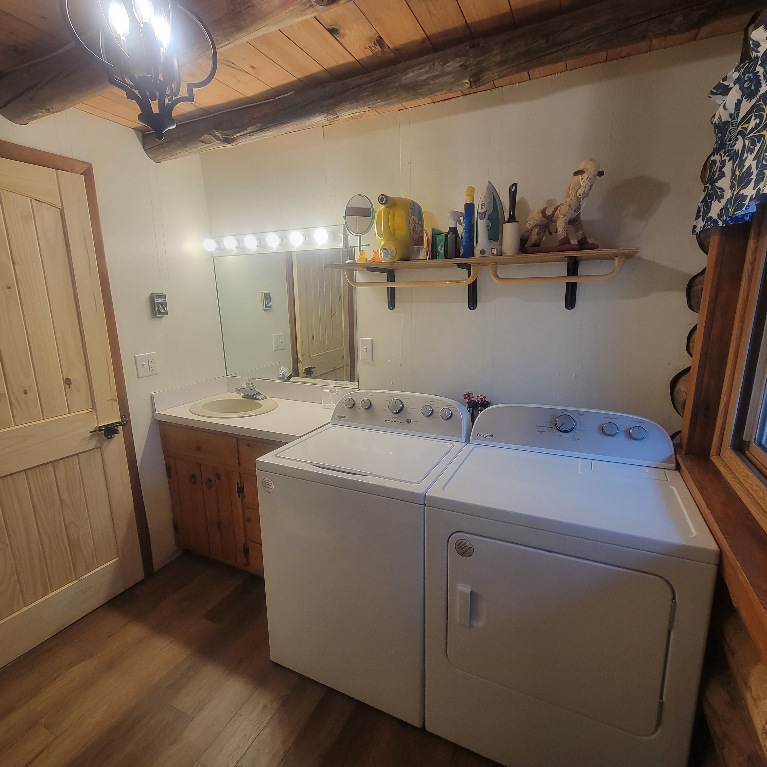 A laundry room with two white Whirlpool washers and dryers, a wooden door, a mirror above the sink, and a shelf with cleaning supplies.