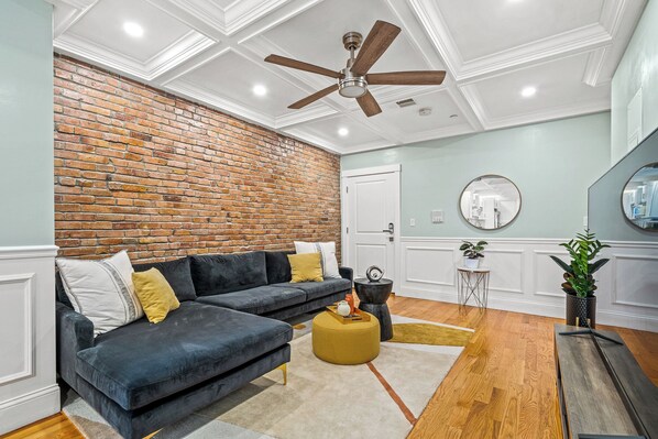 A living room with a dark blue sofa, a round ottoman, a ceiling fan, a brick wall, and a wooden floor.