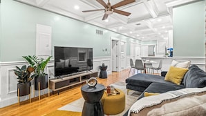 A modern living room with a flat-screen TV, a ceiling fan, and a dining area in the background.