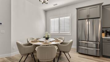 A modern kitchen with a stainless steel refrigerator, a dining area with a round table and four chairs, and a window with blinds.