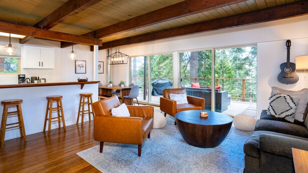 A spacious living area with wooden flooring, a bar area, and large windows offering a view of the outdoors.