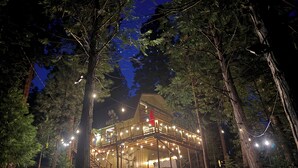 A multi-level wooden cabin with string lights, surrounded by trees.