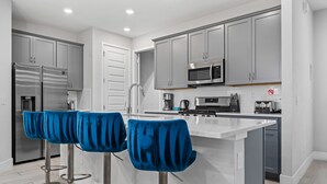 Eine moderne Küche mit grauen Schränken, einem Edelstahlkühlschrank, einer Mikrowelle und einer Kaffeemaschine. Zwei blaue Barhocker mit Metallfüßen ergänzen das Bild.