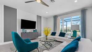 Ein modernes Wohnzimmer mit einem weißen Sofa, einem Türkis-Armlehnstuhl und einem Glastisch. Ein Flachbildfernseher ist an der Wand befestigt, und es gibt ein großes Fenster mit Vorhängen.