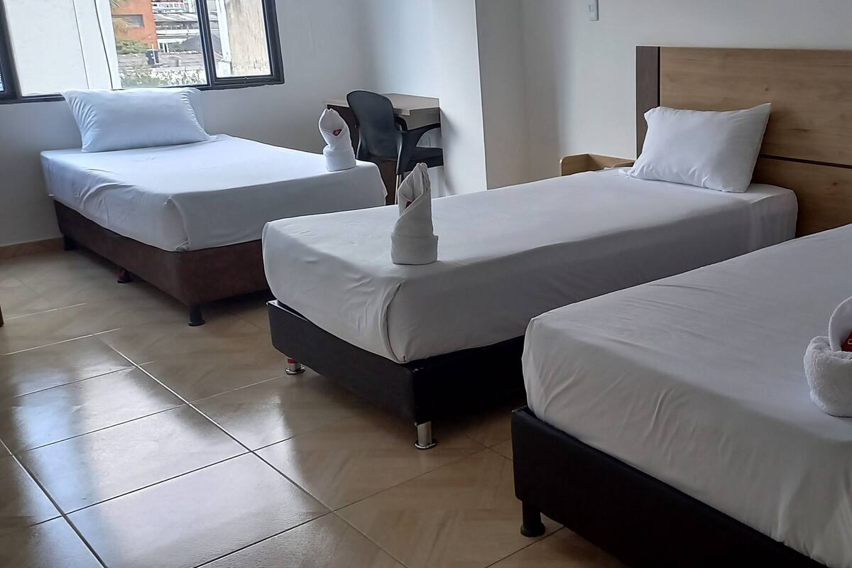 A hotel room with three beds, each with white bedding and pillows. There is a wooden headboard on the right bed. The room has a tiled floor and a window on the left side.