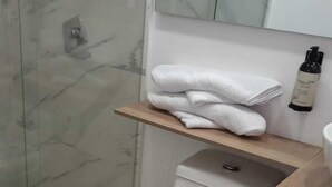 A modern bathroom with a glass shower enclosure, a white toilet, a wooden vanity, and a shelf with folded towels. - Hotel Boutique FC (Bogotá)