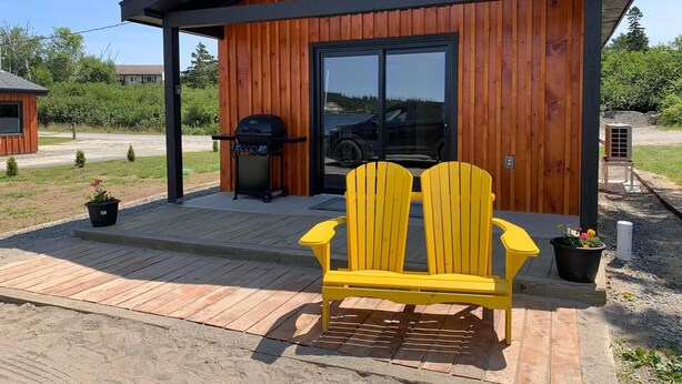 Een moderne houten blokhut met een grote schuifdeur, een barbecue en twee gele Adirondack-stoelen op een houten terras.
