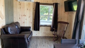 Een kamer met houten lambrisering, een leren bank, een schommelstoel, een aan de muur bevestigde televisie en een raam met gordijnen.