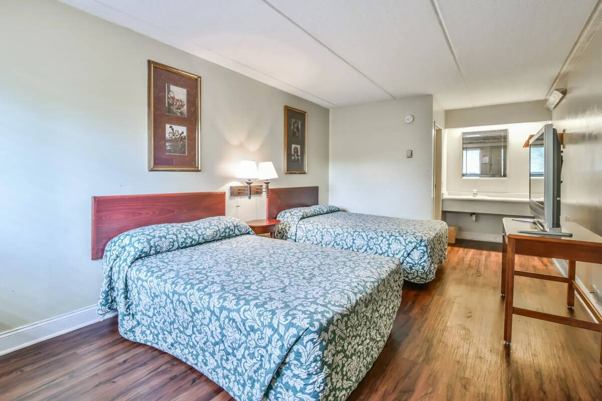A hotel room with two beds, wooden headboard, framed pictures on the wall, a bedside table with lamps, and a wooden desk with a mirror.