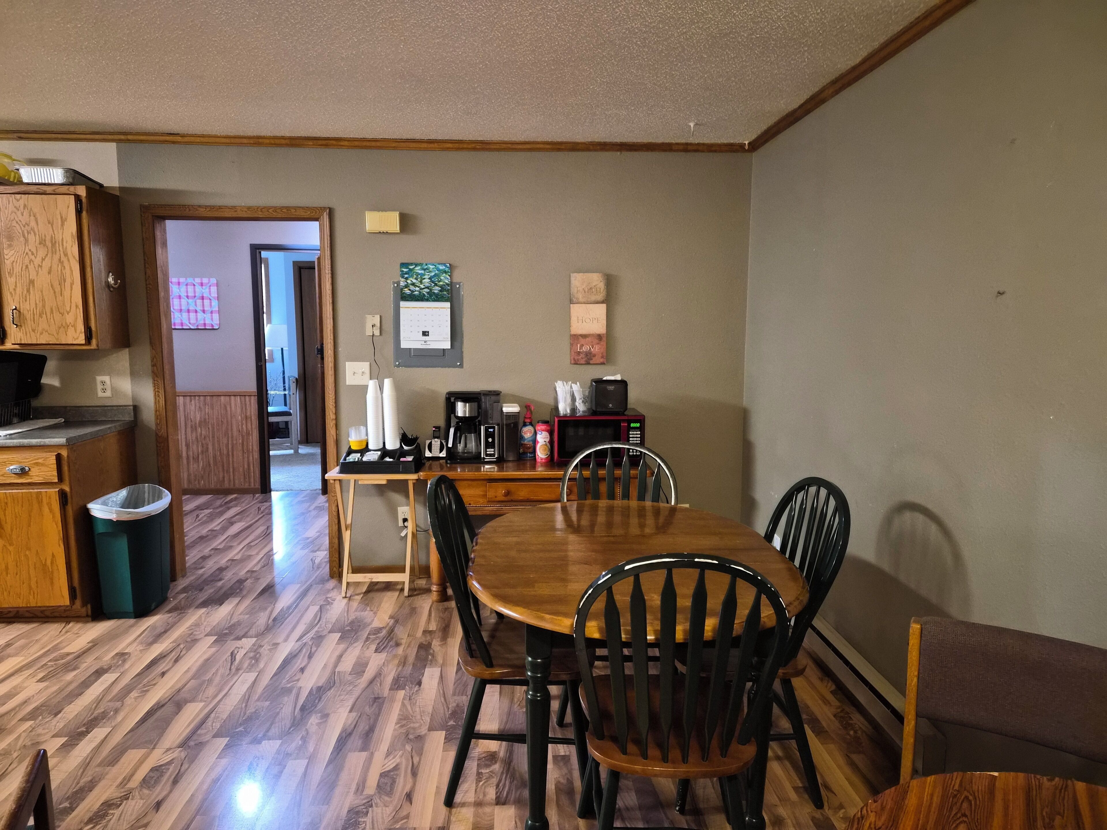 A dining area with a wooden table, four chairs, a trash bin, a microwave, a coffee maker, and a calendar on the wall.