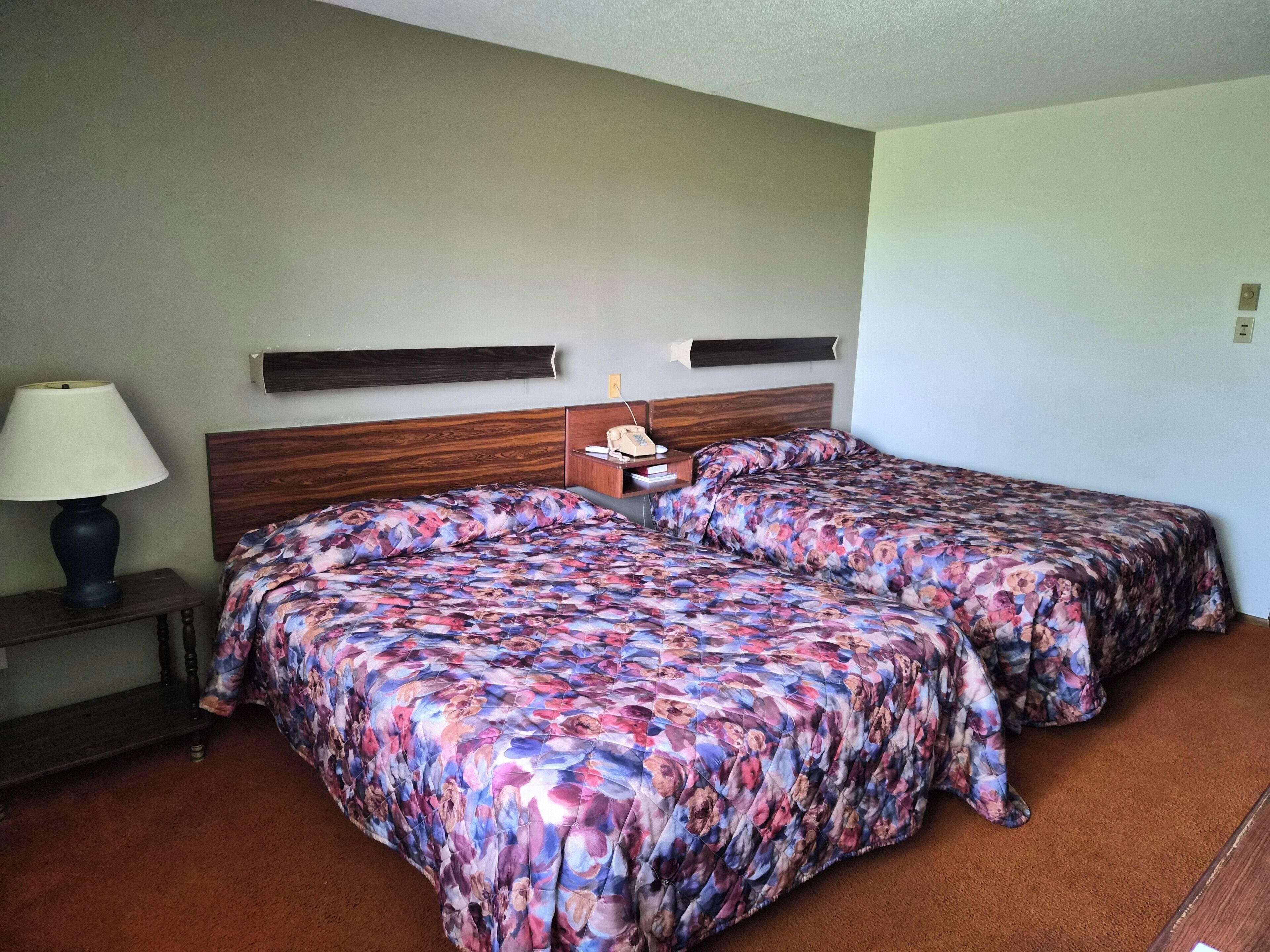 A hotel room with two beds, a nightstand, a lamp, and a wall-mounted heater.