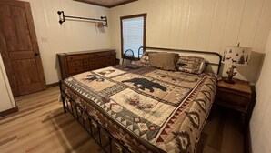 Un dormitorio con techo de madera, una cama con colcha estampada, una mesita de noche con lámpara y una ventana con persianas.