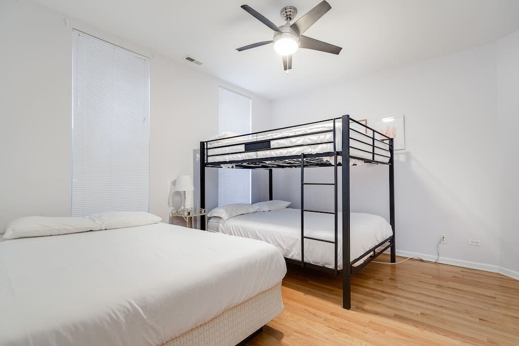 A room with a bunk bed, a single bed, a ceiling fan, and a window with blinds.