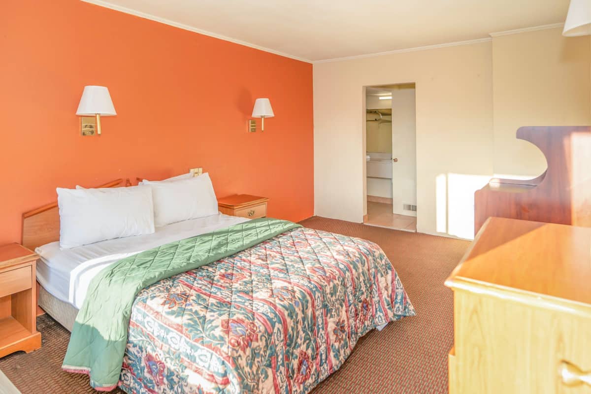 A hotel room with a bed, two bedside lamps, a wooden nightstand, a dresser, and a door leading to a bathroom.