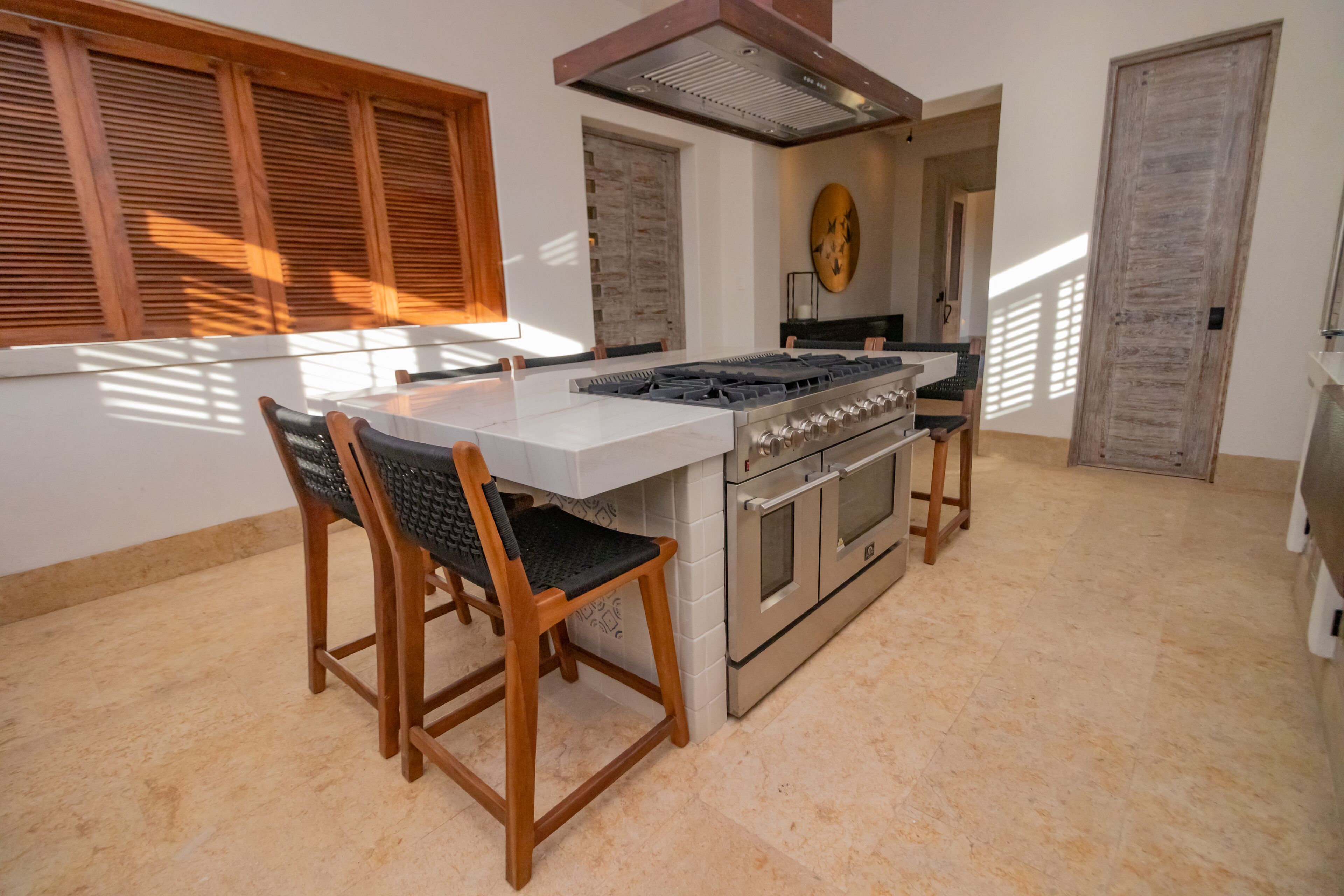 A modern kitchen with a central island, wooden chairs, a stove, and a large window with blinds.