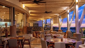 Un restaurant spacieux avec des tables et des chaises en bois, de grandes fenêtres offrant une vue sur le coucher du soleil, et un intérieur chaleureux avec des poutres apparentes et des ventilateurs de plafond.