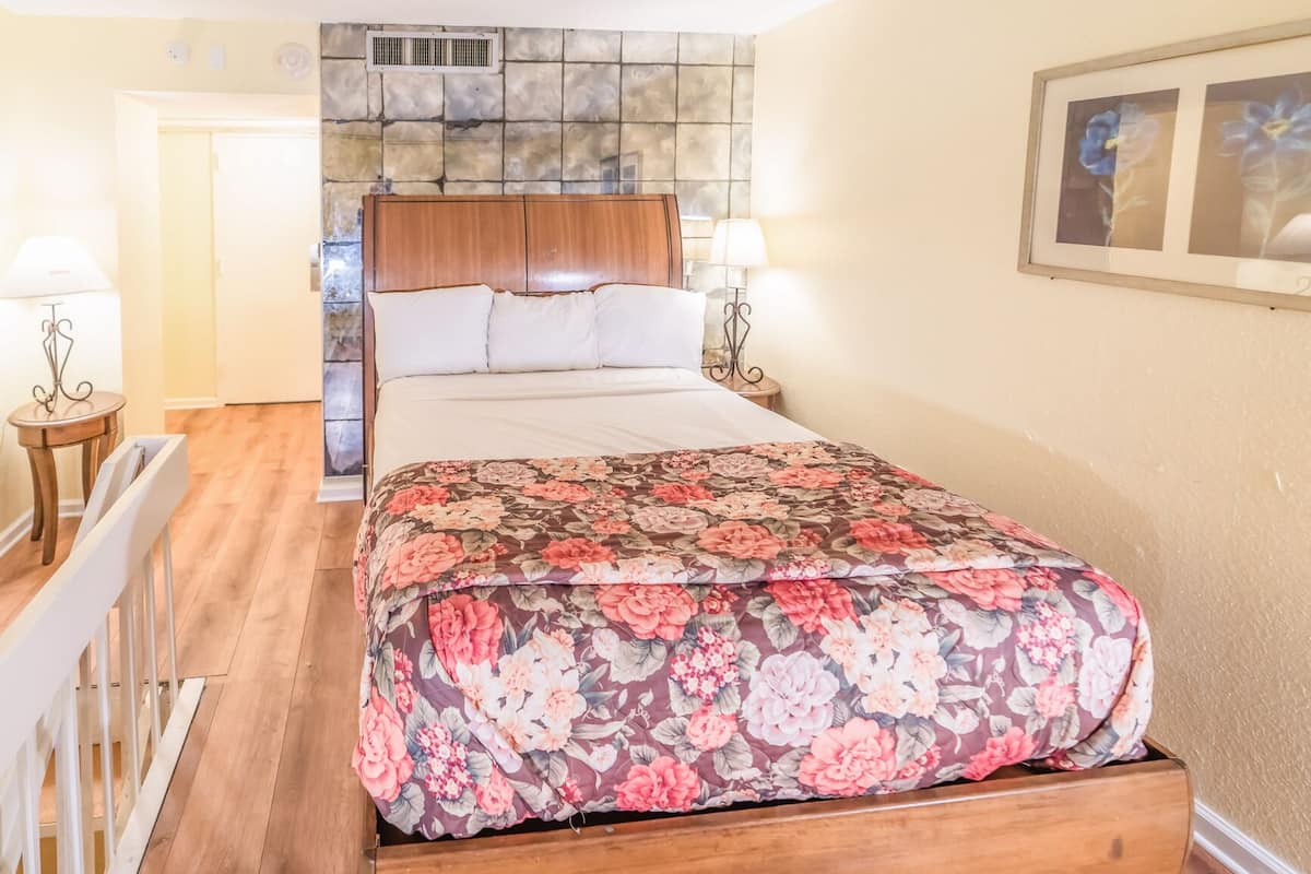 A bedroom with a wooden bed, floral bedspread, two bedside lamps, a stone wall, and a framed picture on the wall.