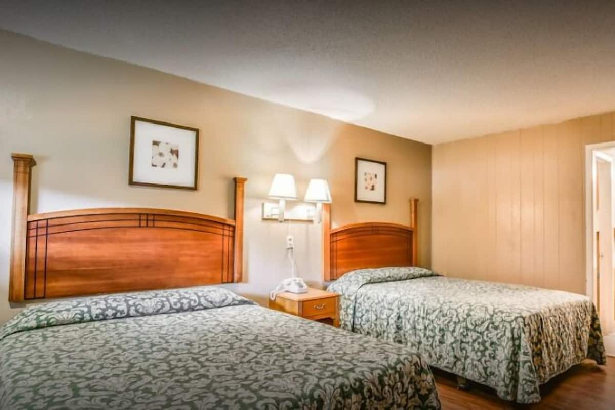 A hotel room with two beds, wooden headboards, a bedside table with a lamp, and a bathroom visible through an open door.