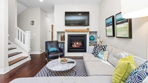 A modern living room with a fireplace, a mounted TV, and a staircase. - Modern Cottage "M" Luxury home w/hot tub, community pool-walk to town & beach (New Buffalo)