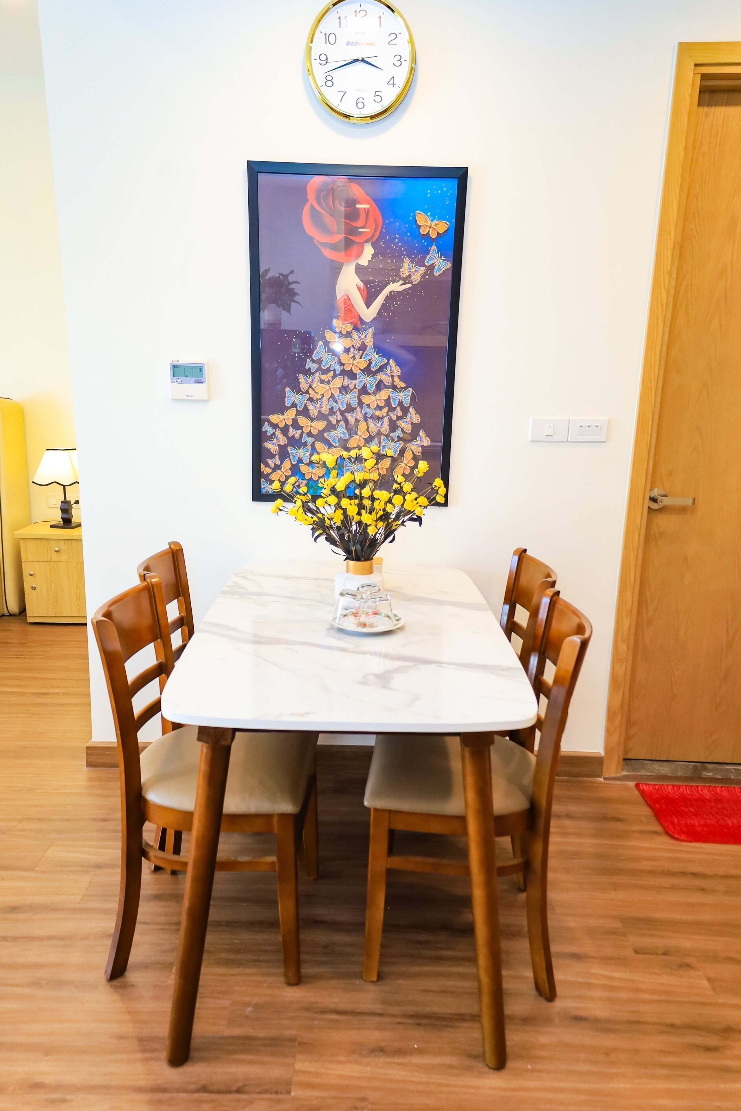 Ein Essbereich mit einem weißen Tisch, zwei Holzstühlen, einer Vase mit gelben Blumen, einer Uhr und einem gerahmten Kunstwerk an der Wand.