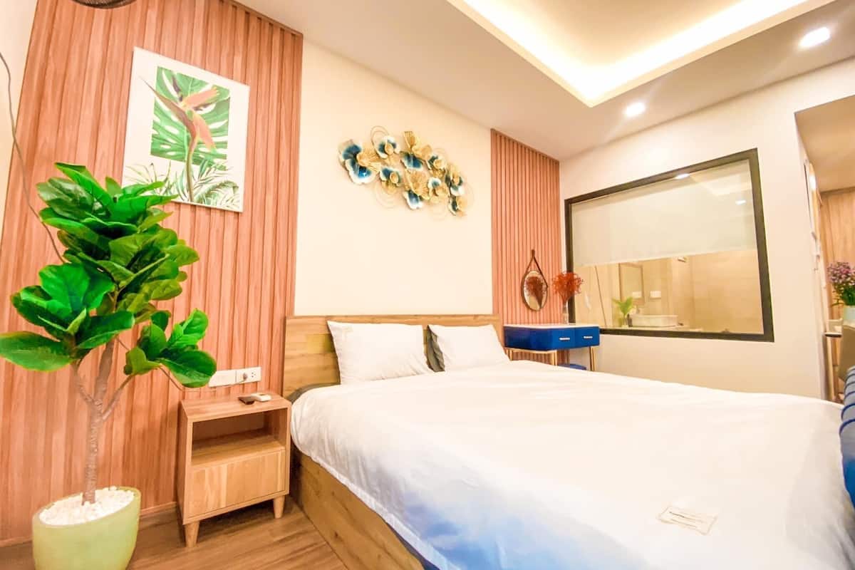 A modern bedroom with a large bed, wooden headboard, a small bedside table, a nightstand, a potted plant, a wall-mounted mirror, and a decorative wall piece.