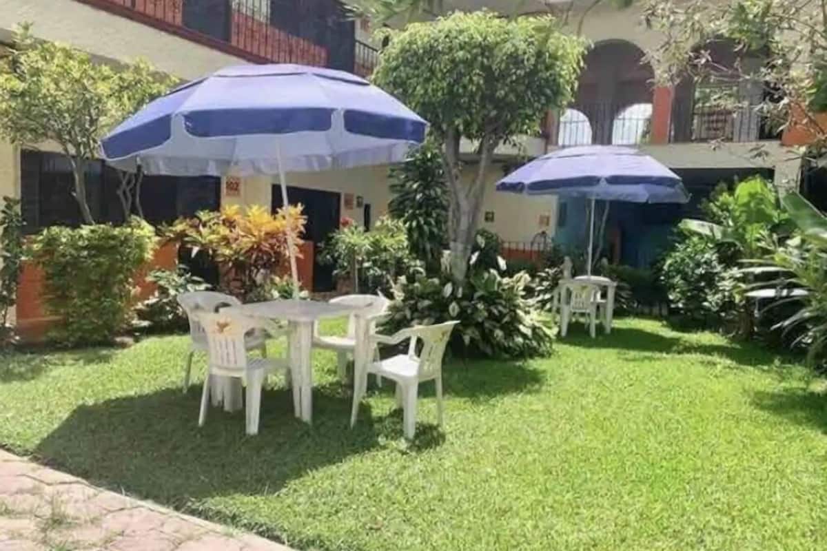 Un patio con una mesa y sillas, rodeado de vegetación y cubierto por un paraguas azul.