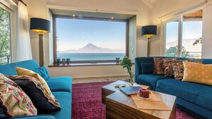 Una sala de estar con dos sofás azules, una mesa de centro de madera y un ventanal con vistas a la montaña y al mar.