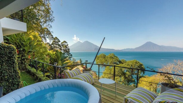 Una terraza exterior con jacuzzi, zona de estar y vistas a una montaña y un cuerpo de agua.