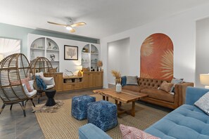 A modern living room with wicker chairs, a sofa, ottomans, and a wooden coffee table. There are shelves with decorative items and a large abstract painting on the wall.