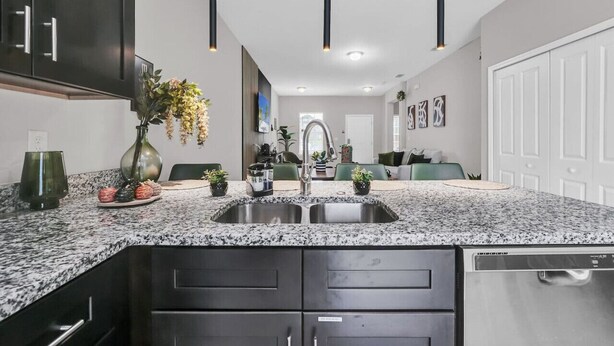 A modern kitchen with granite countertops, stainless steel sink, and black cabinetry. There are pendant lights, a vase with flowers, and a dishwasher. - Uniquely Chic home near Disney! (Kissimmee)