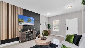 A modern living room with a flat-screen TV, a round coffee table, a sofa with green cushions, a green armchair, and a potted plant. - Uniquely Chic home near Disney! (Kissimmee)