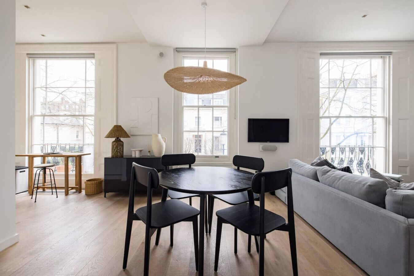 A modern living room with a round dining table, black chairs, a grey sofa, and a TV mounted on the wall.