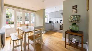 Una cocina moderna con armarios blancos, una zona de comedor con una mesa y sillas de madera, y un jarrón verde sobre una mesita auxiliar.