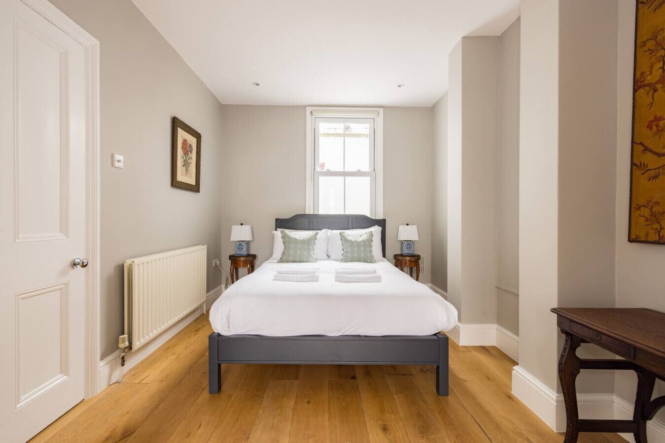 A bedroom with a bed, two bedside tables, a radiator, a window, and a painting on the wall.
