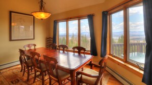 A dining room with a wooden table and chairs, large windows with curtains, and a hanging light fixture.