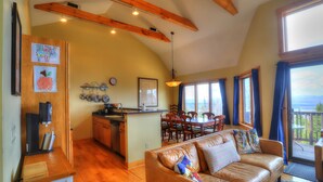 A cozy living room with a leather couch, wooden beams, a kitchen area, and large windows offering a view.