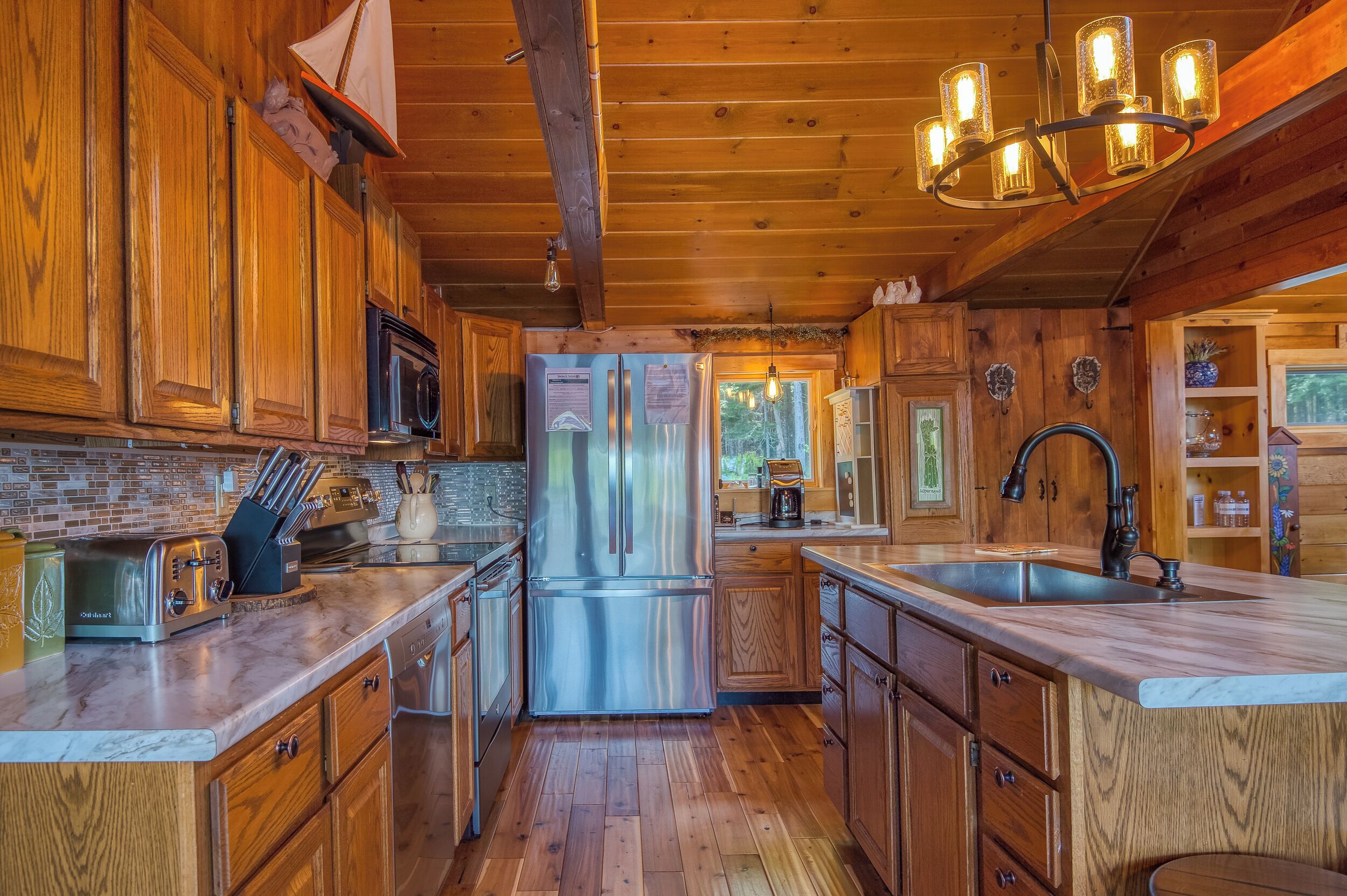 Une cuisine rustique aménagée dans une cabane en bois, avec un réfrigérateur en acier inoxydable, des placards en bois, un plan de travail en marbre et un luminaire suspendu.