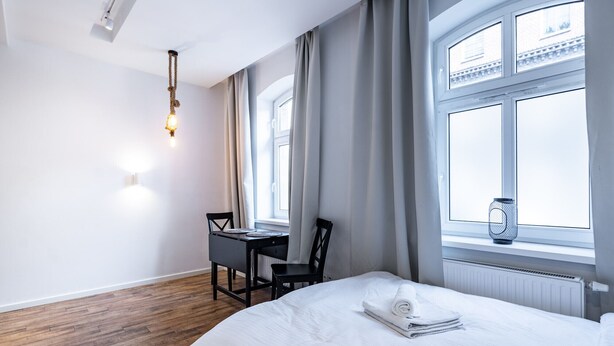 A hotel room with a bed, a desk, a chair, a window with curtains, and a hanging light fixture. - Janasa Comfort Studio (Katowice)