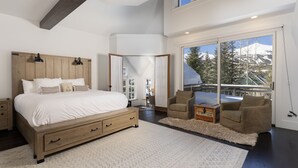 Un dormitorio con una cama grande, cabecera de madera, dos lámparas de noche, una mesita de noche, un aparador, una alfombra mullida, dos sillones, una pequeña mesa y un ventanal con vistas a montañas nevadas.