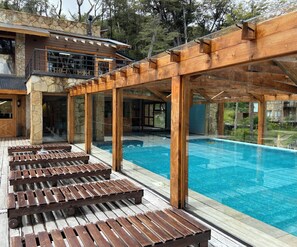 A modern outdoor pool area with wooden loungers and a stone building in the background. - El Condor Pasa VLA  (Villa La Angostura)